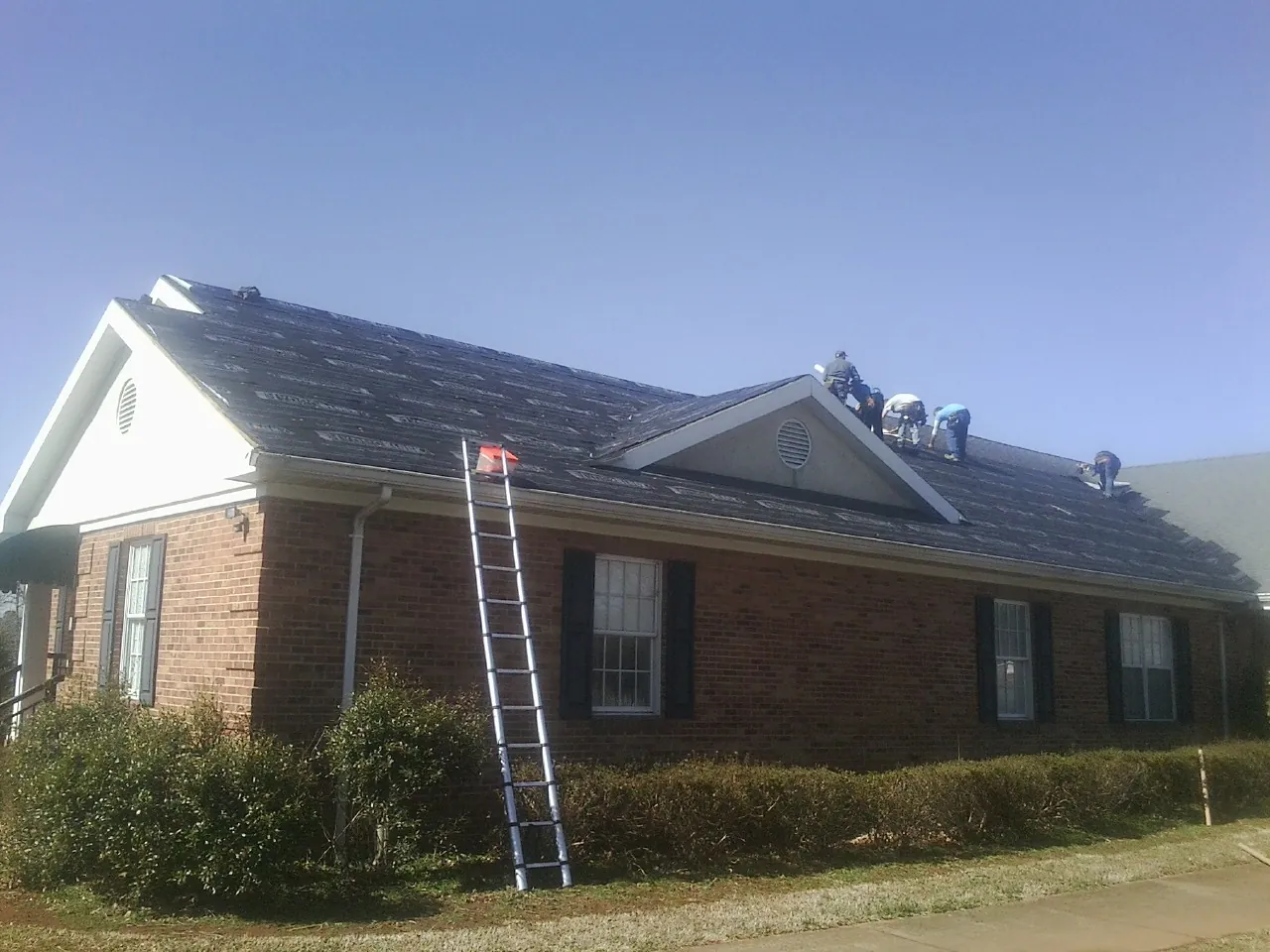 Crew tearing shingles off a brick ranch roof with a ladder set — Emergency Roof Repair work by our crew in Candor