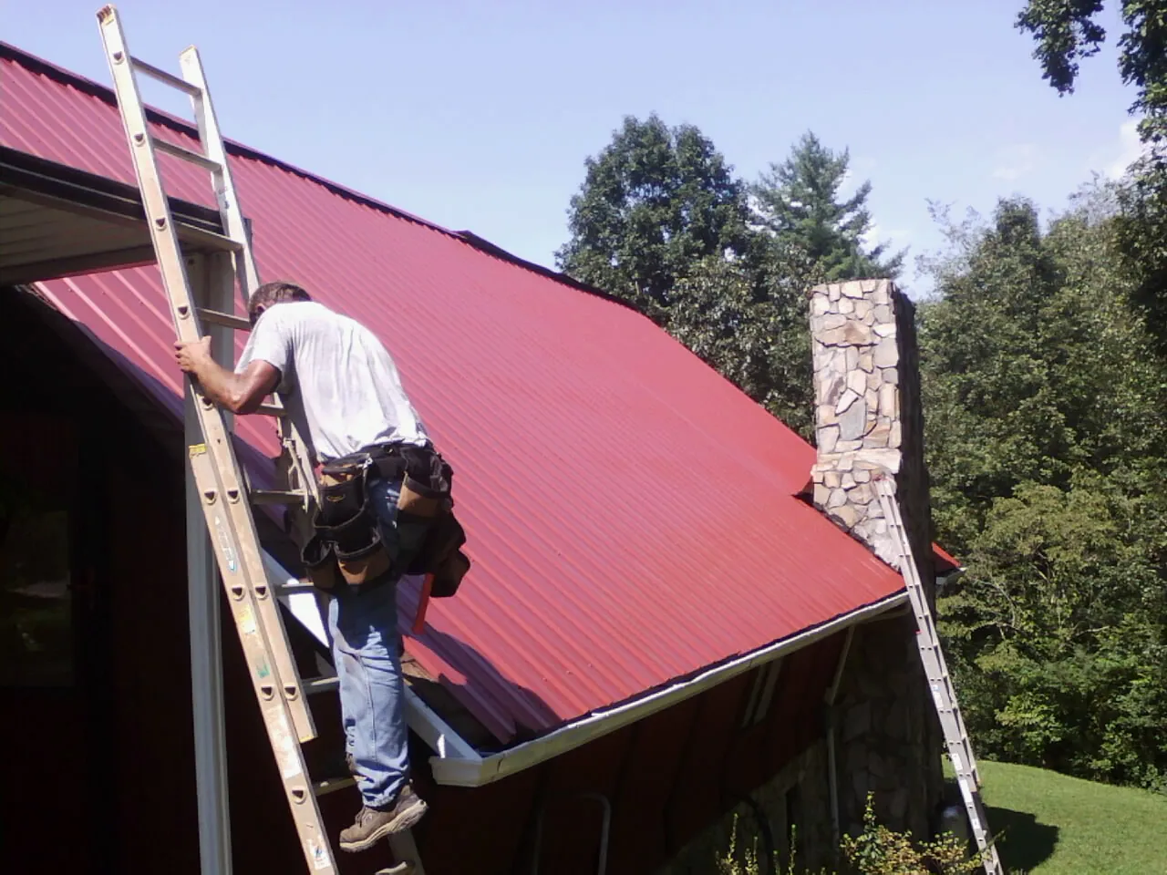 Roofer on a ladder beside a red metal roof and stone chimney — Soffit & Fascia Repair by Roofing Brothers in Candor